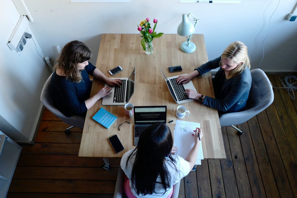 Three women sit around a timber table with their laptops. Co-working or working from cafes is a way of supporting remote working in 2023