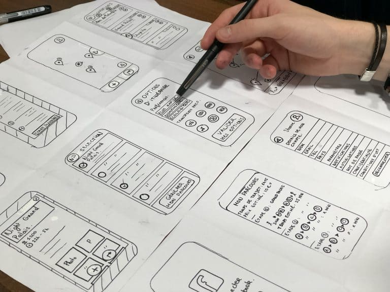 A large sheet of paper on a desk, a hand holding a marker on the right. Handdrawn smartphone icons are in rows, each with design options