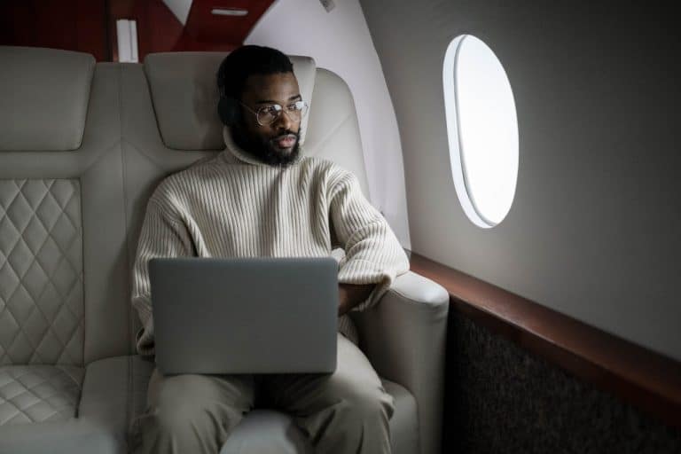 A man sits in a first class seat on a plane, looking out the window, accessing onedrive sharepoint teams offline via a laptop balanced on his lap.